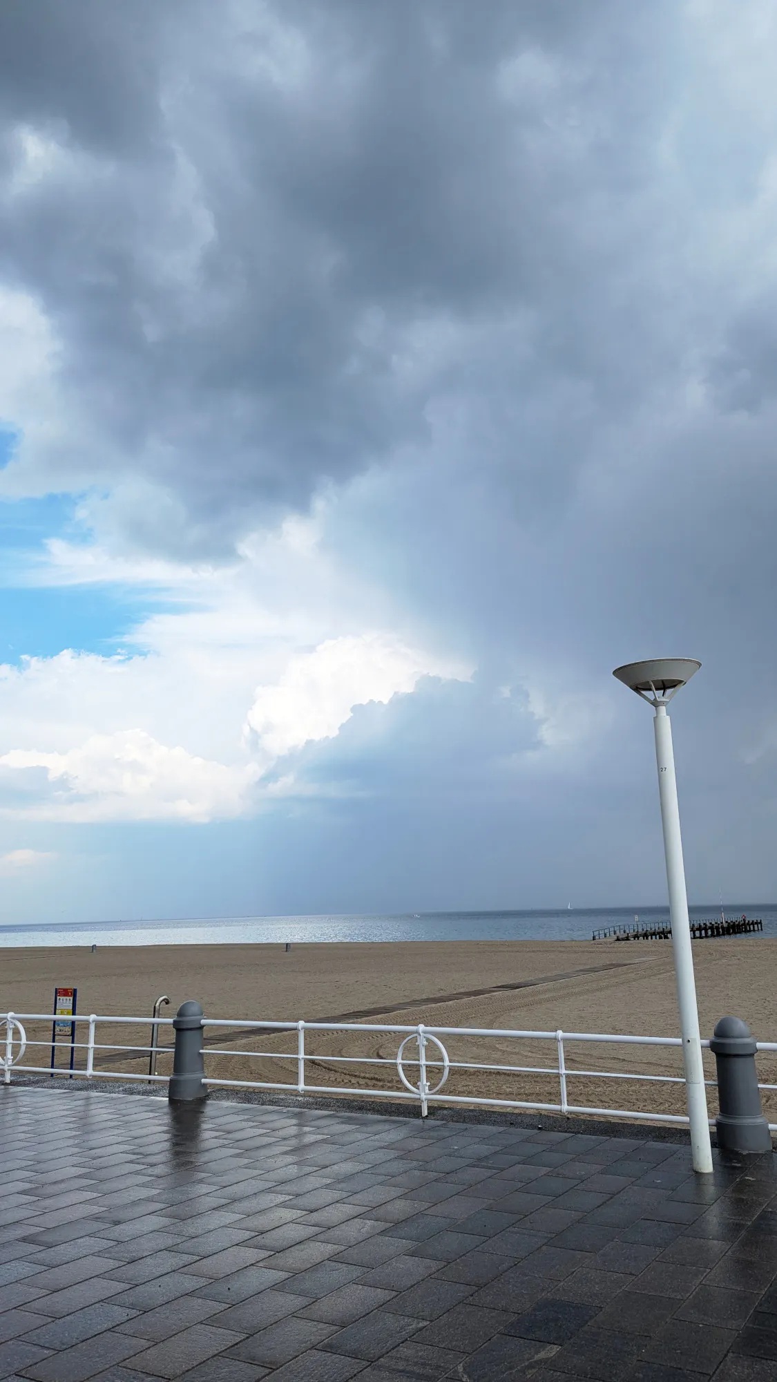 Regennasses Pflaster einer Strandpromenade, dahinter Strand, dahinter Meer, über allem dunkelgraue und leuchtend weiße Wolken und ein Fleck blauer Himmel.