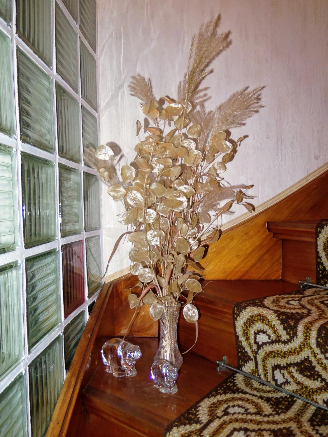 Eine Treppe aus Holz, darauf ein grob gewebter Läufer braun in braun, strenge Trockenblumen in einer gläsernen Vase, davor zwei kleine Bären aus Glas. Zum Abschluss Glasbausteine.