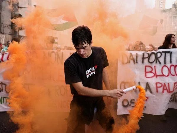 Bild vom Gernalstreik in Italien, 1 junger Mensch vor einer Reihe von Protestierenden, die Transparente halten, die Person im Vordergrund trägt ein schwarzes Tshirt und hält einen Rauchtopf, aus dem oranger Rauch steigt.