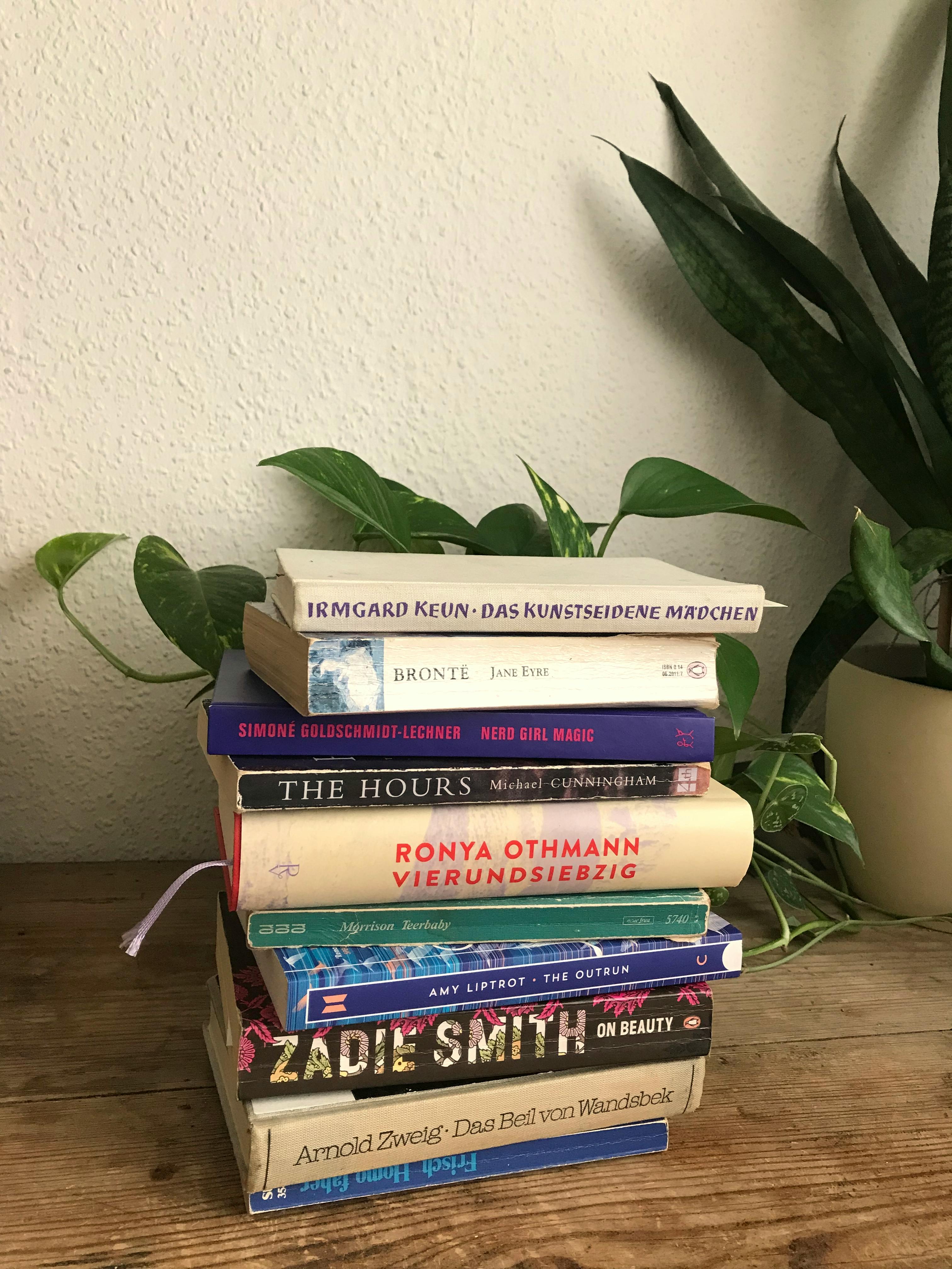 bücherstapel auf holztisch, zimmerpflanzen im hintergrund, buchrücken von oben: das kunstseidene mädchen, jane eyre, nerd girl magic, the hours, vierundsiebzig, teerbaby, the outrun, on beauty, das beil von wandsbek. zuunterst und verkehrt herum: homo faber. / staple of books on wooden table, house plants in the background, book spines from above: das kunstseidene mädchen, jane eyre, nerd girl magic, the hours, vierundsiebzig, teerbaby, the outrun, on beauty, das beil von wandsbek. at the bottom and upside down: homo faber.