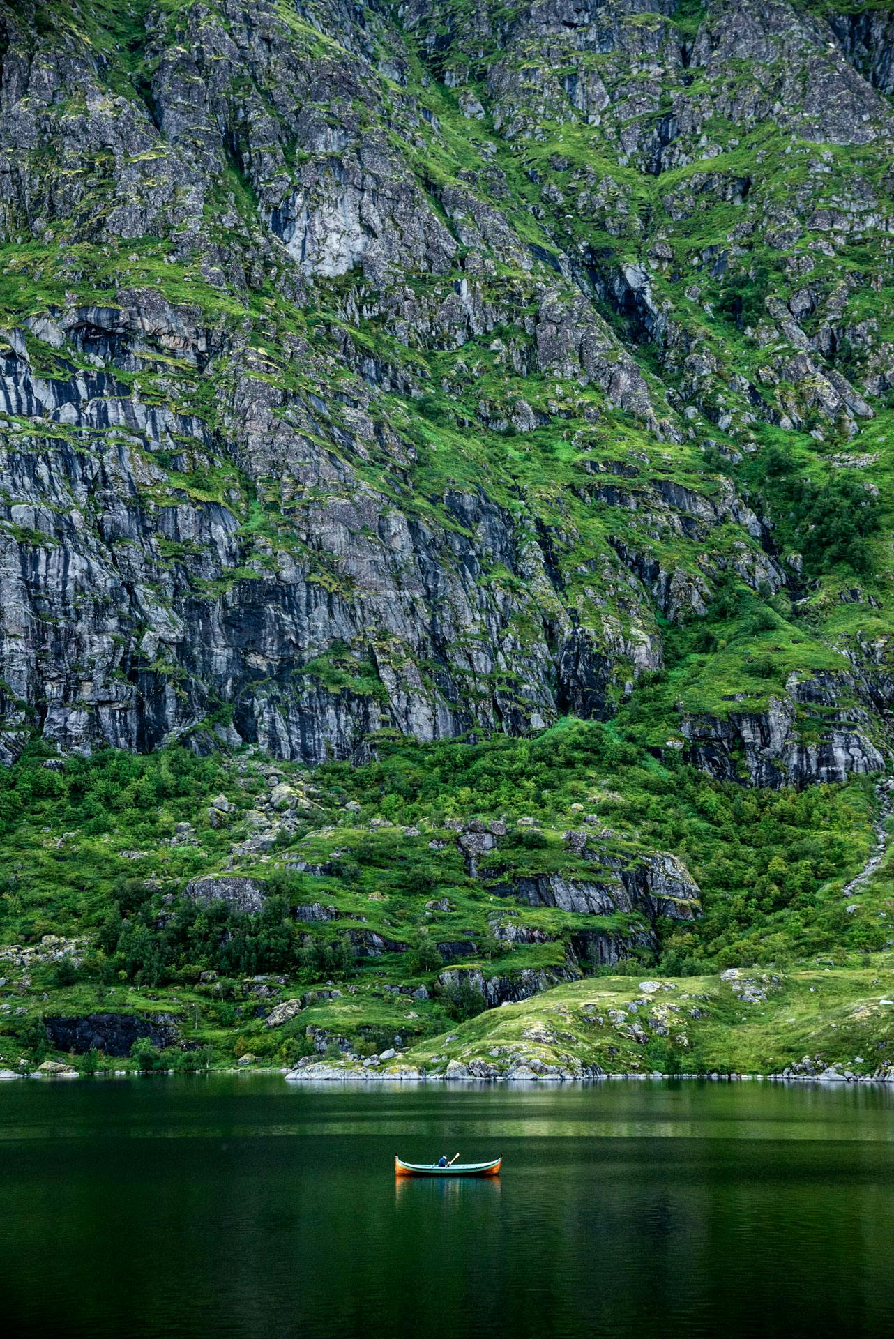 Une petite barque typique des îles Lofoten, sur un lac d'un vert émeraude aux eaux très calmes. Derrière elle se dressent des falaises rocheuses qui prennent tout le cadre de l'image.