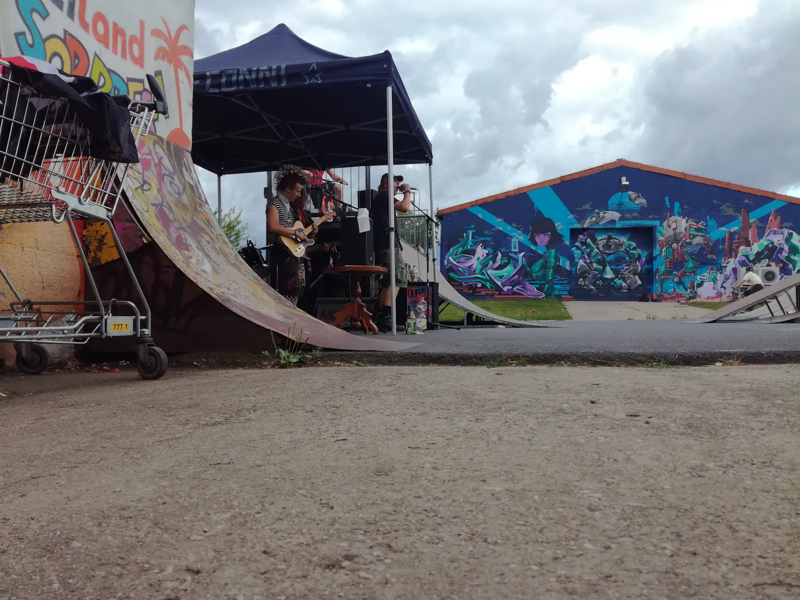 Punkband auf weitläufigem Gelände. Zwischen Skateanlagen, vorne links ein Einkaufswagen. Zuschauer sind nicht zu sehen.