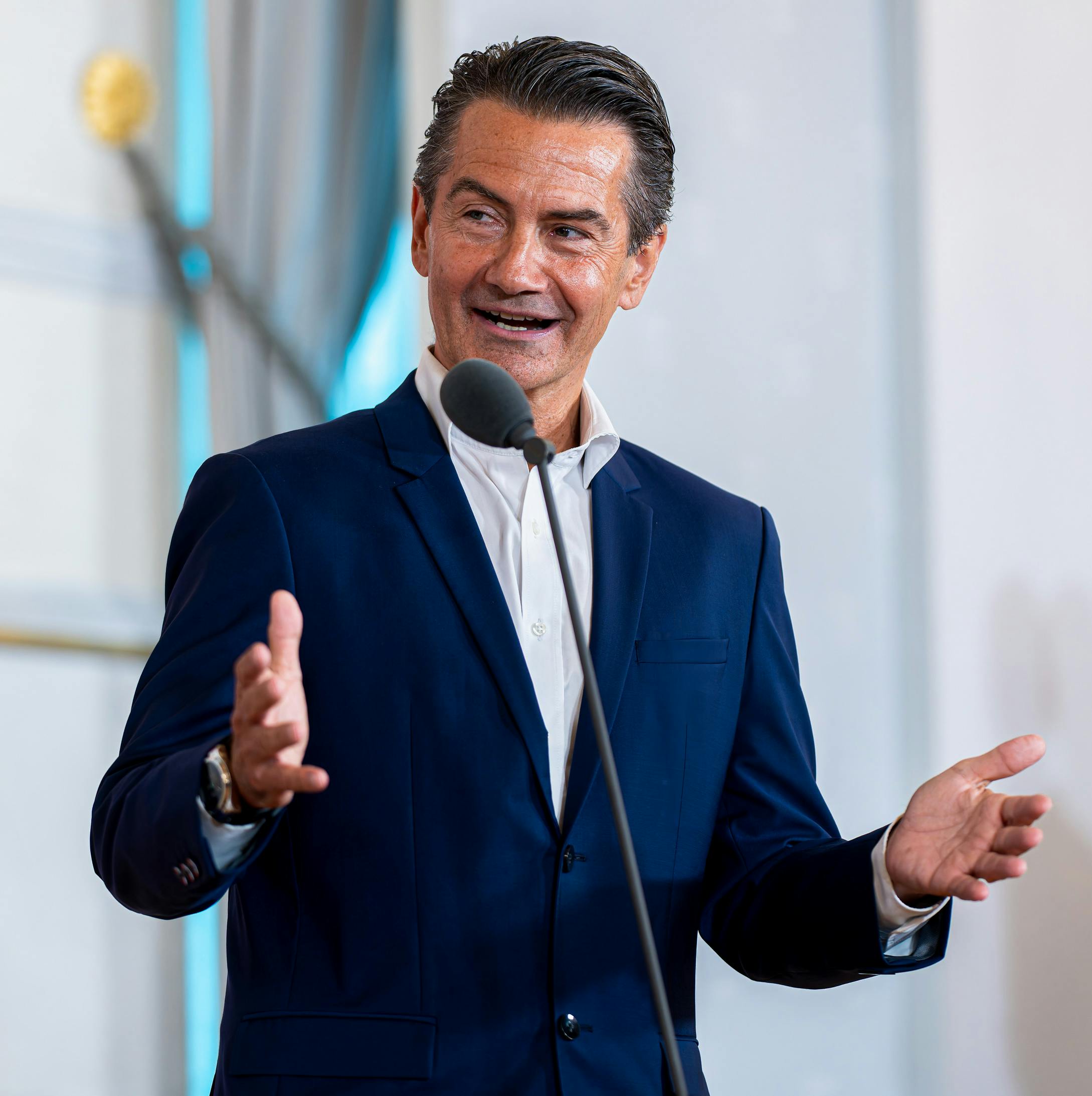 Das Bild zeigt Roland Weißmann in einem dunkelblauen Sakko, der an einem Mikrofon spricht und dabei beide Hände hebt.