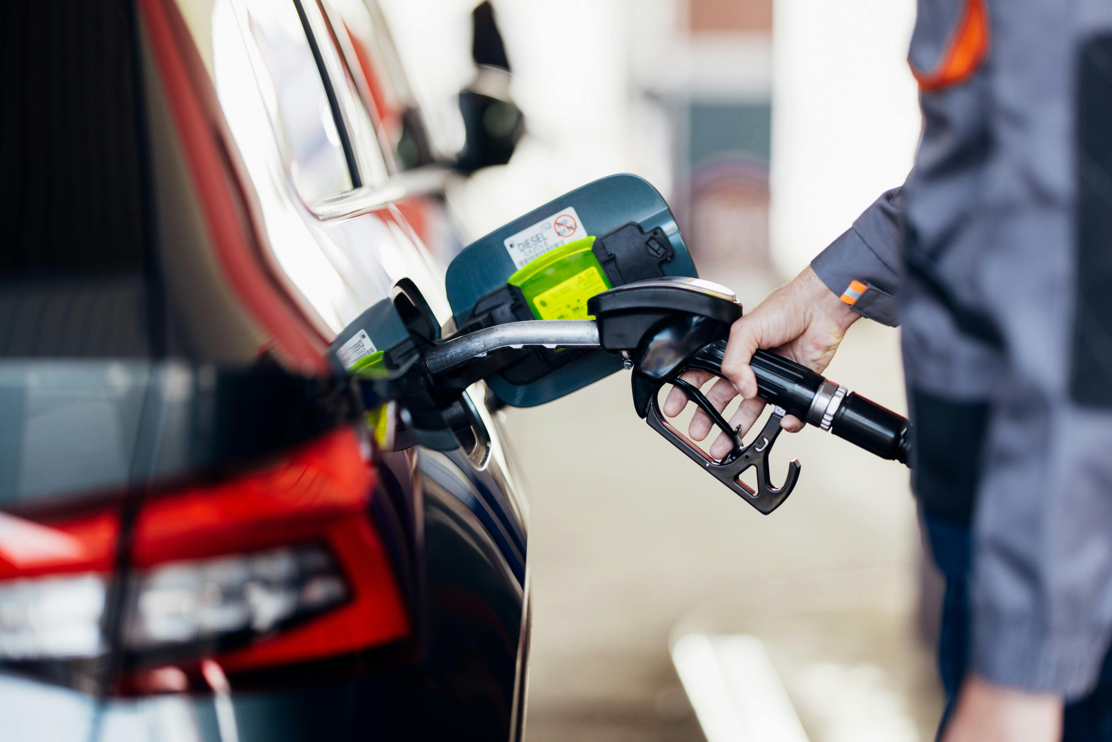 Das Bild zeigt eine Hand, die eine Zapfpistole hält, während an einer Tankstelle ein dunkler SUV getankt wird. Im Hintergrund ist die rote Rückleuchte des Autos zu sehen.