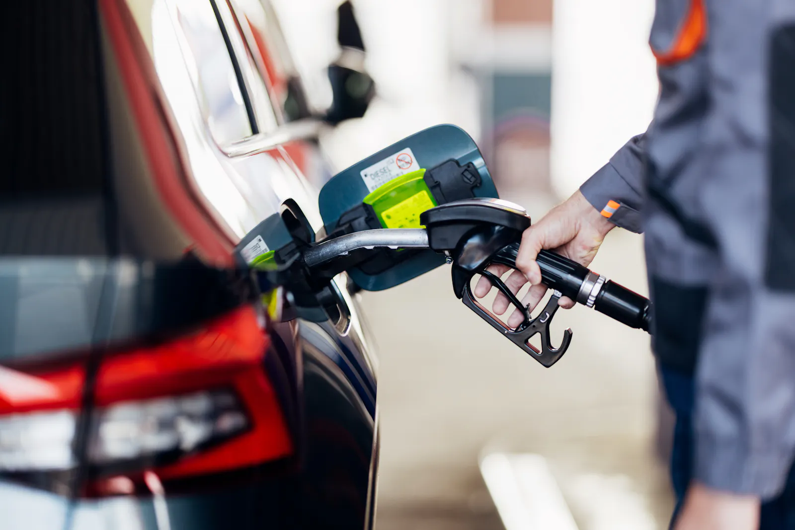 Das Bild zeigt eine Hand, die eine Zapfpistole hält, während an einer Tankstelle ein dunkler SUV getankt wird. Im Hintergrund ist die rote Rückleuchte des Autos zu sehen.