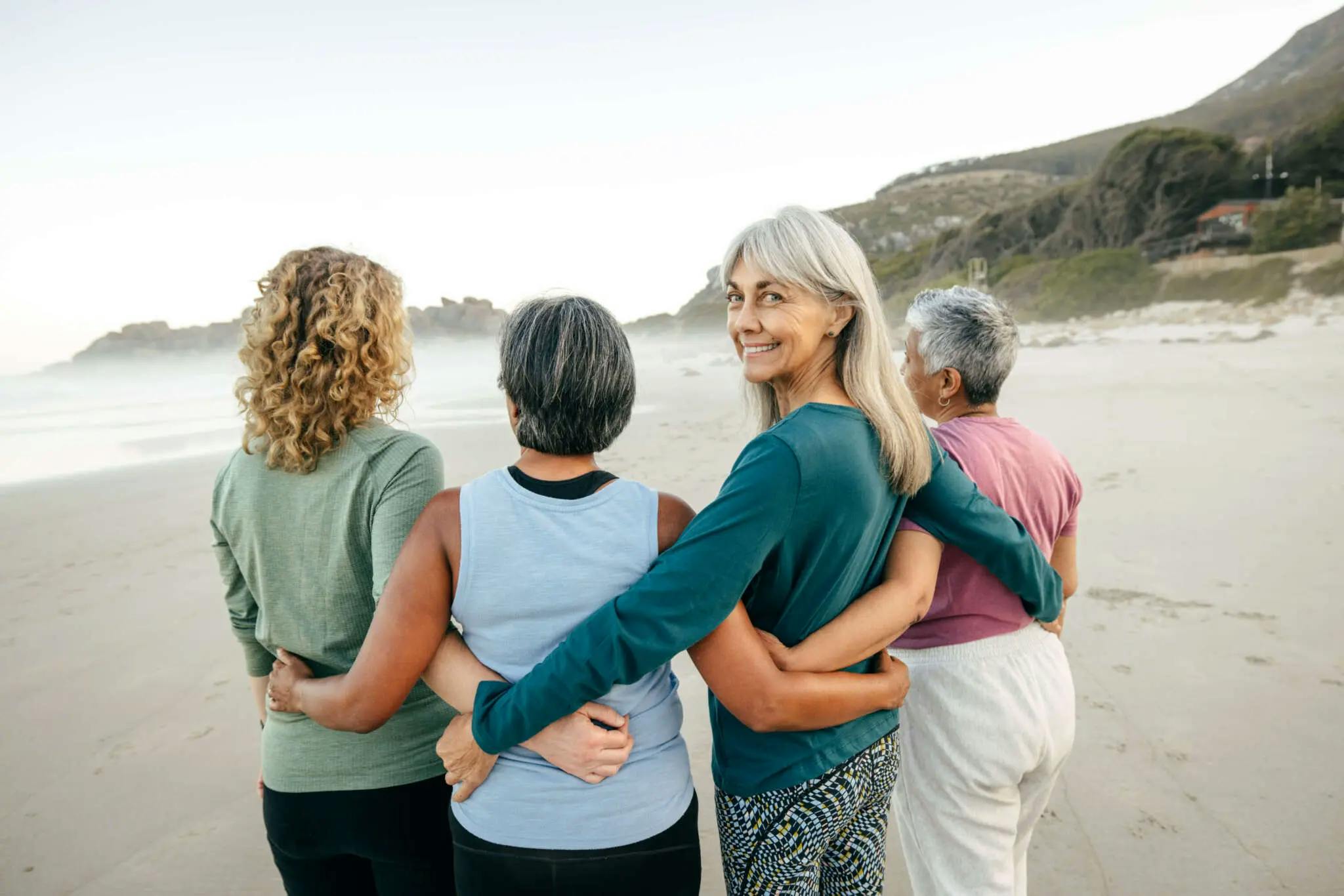 4 Frauen mittleren Alters stehen sich umarmend in einer Reihe, mit dem Rücken zur Kamera, an einem Strand. Eine Frau dreht sich um und lächelt in die Kamera