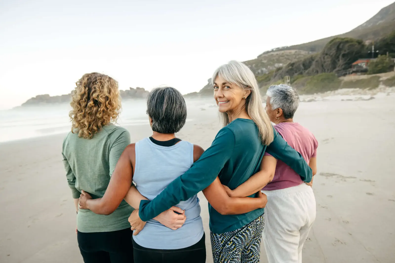 4 Frauen mittleren Alters stehen sich umarmend in einer Reihe, mit dem Rücken zur Kamera, an einem Strand. Eine Frau dreht sich um und lächelt in die Kamera