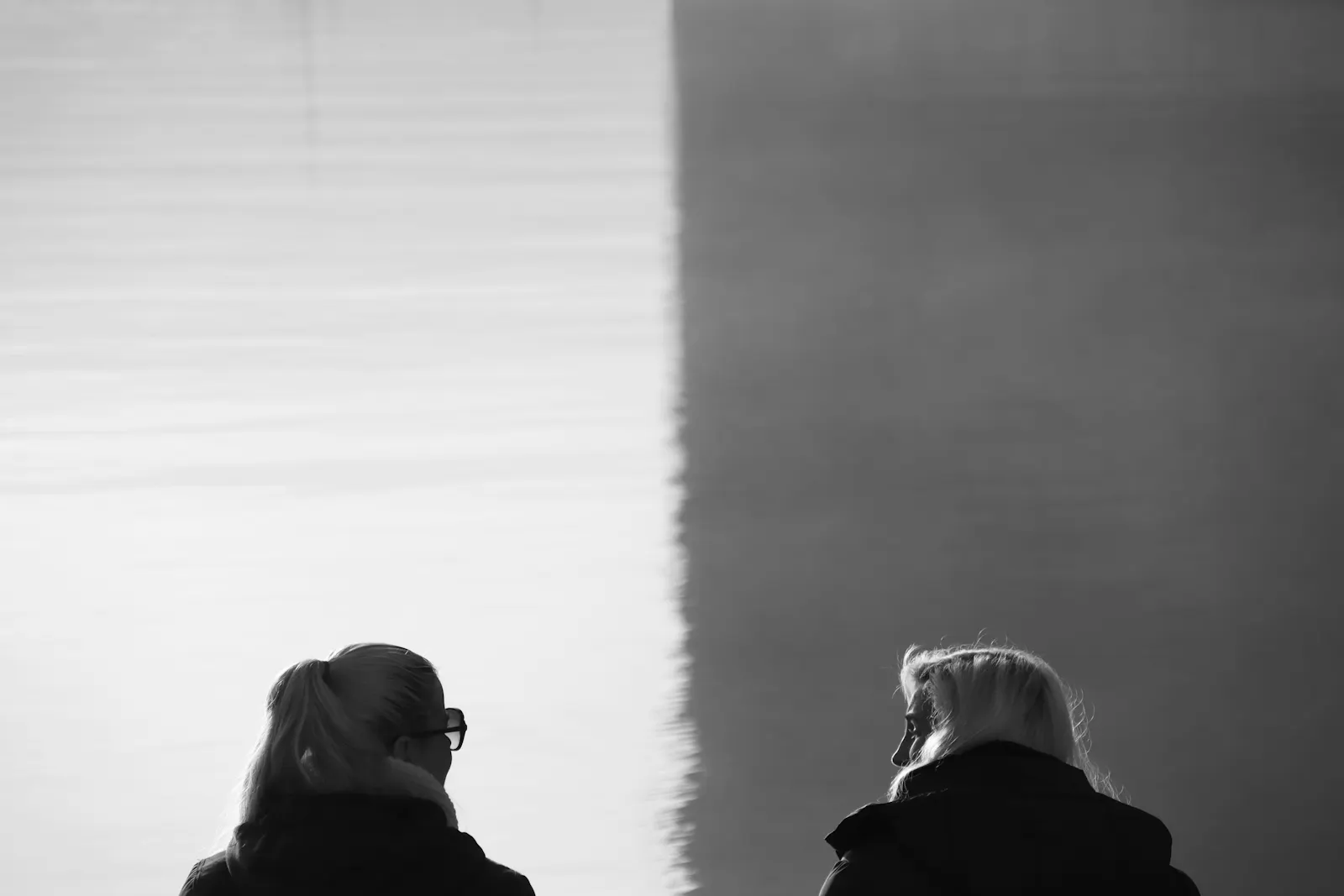 Schwarz-Weiß: Zwei Personen vor einer Wasserfläche. DUrch Schattenwurf mittig rechts heller, links dunkler. Die Personen blicken sich an.