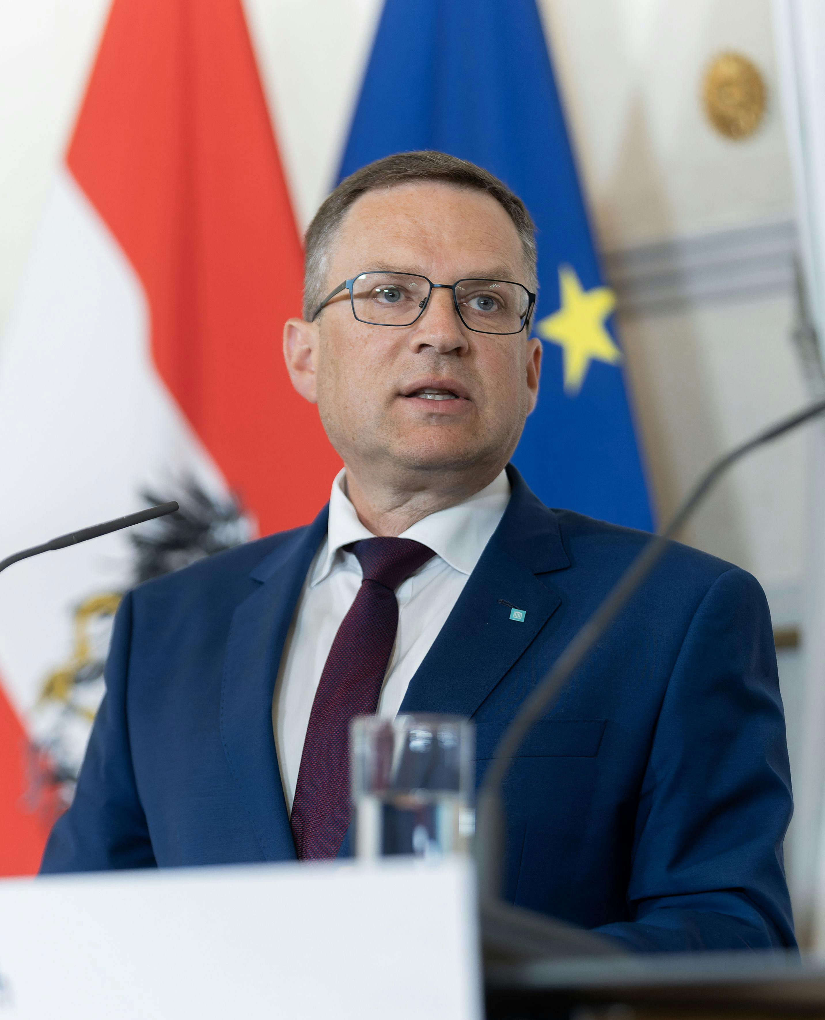 Das Bild zeigt den ÖVP-Politiker August Wöginger. Er trägt einen blauen Anzug und spricht vor der österreichischen Flagge und der EU-Flagge.