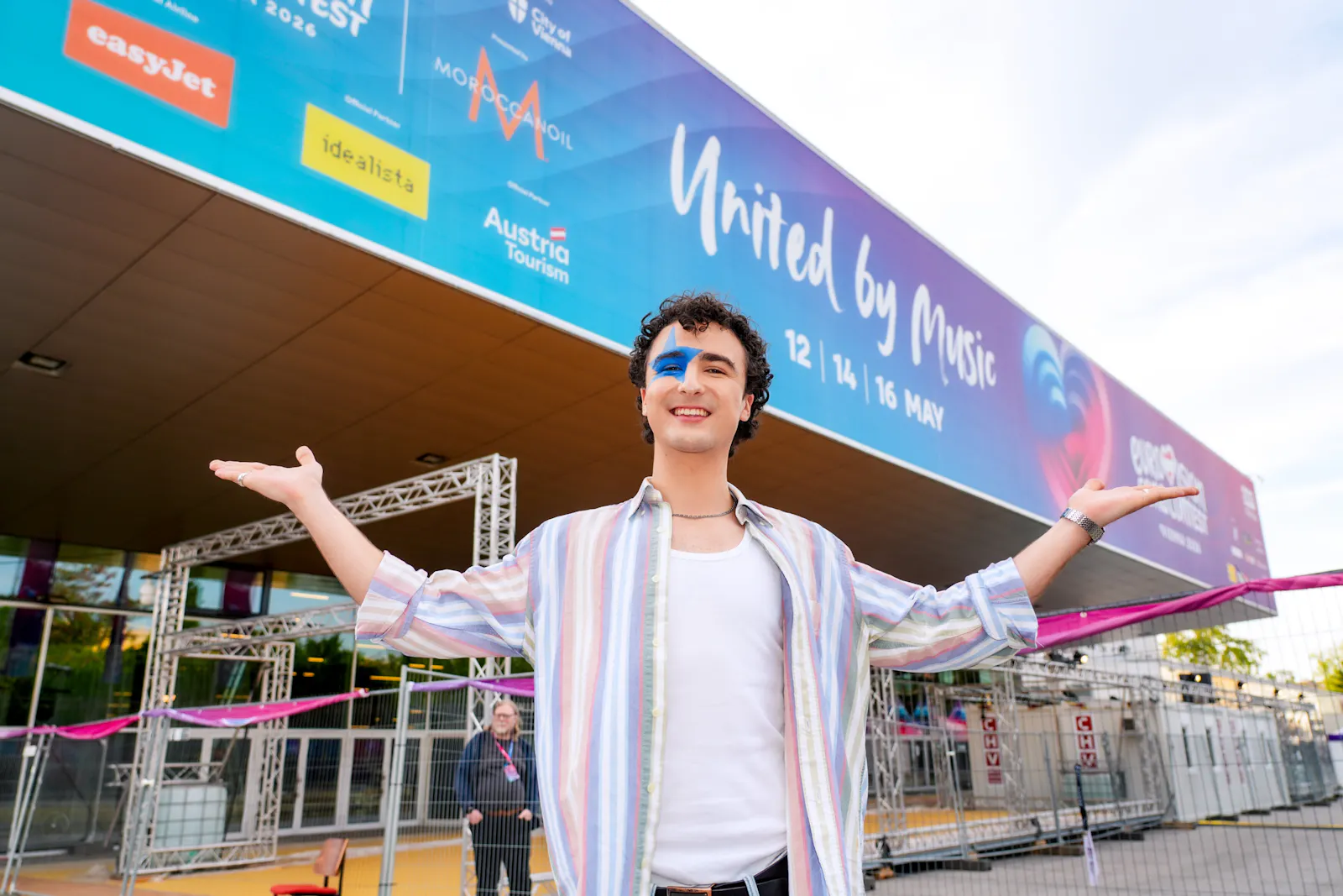 Das Bild zeigt den Sänger Cosmos, der mit ausgebreiteten Armen und einem Lächeln vor der Wiener Stadthalle steht. Im Hintergrund ist ein großes Banner mit der Aufschrift „United by Music" und den Daten 12, 14 und 16 Mai zu sehen.