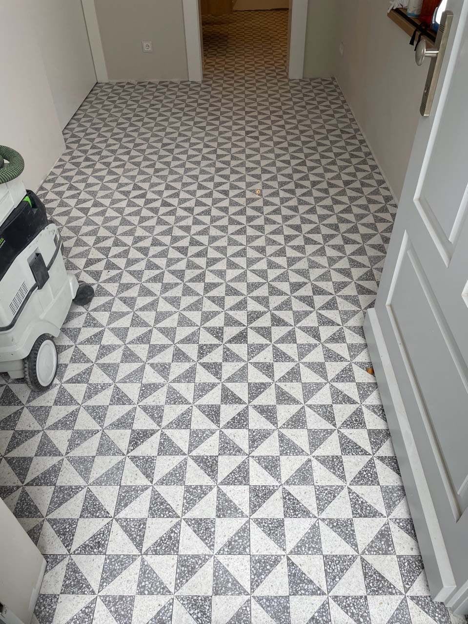 Foto Hausflur hinter geöffneter Haustür mit neu verlegten Mosaikfliesen in grau-weißem Muster.