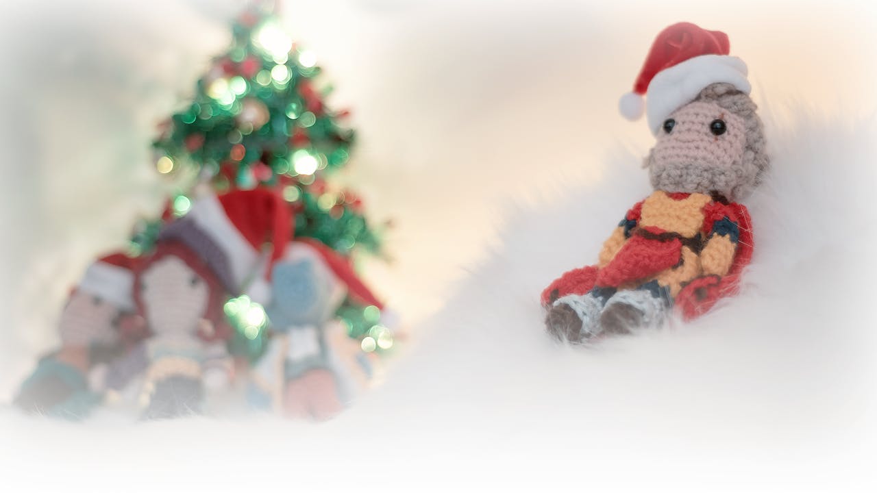 Ein Foto von einem gehäkelten Flynt, der auf weißem Fell sitzt und eine kleine Weihnachtsmütze trägt. Im Hintergrund sitzen Peren, Elaynie und Medrash vor einem kleinen Weihnachtsbaum.
