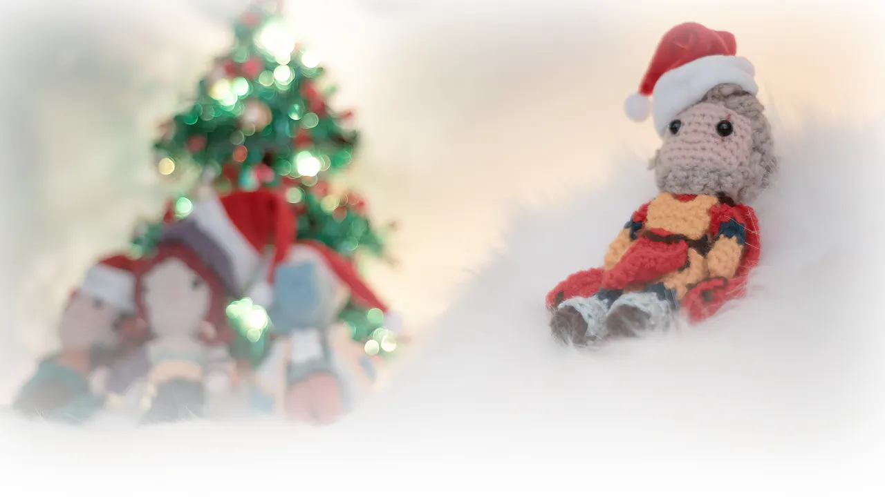 Ein Foto von einem gehäkelten Flynt, der auf weißem Fell sitzt und eine kleine Weihnachtsmütze trägt. Im Hintergrund sitzen Peren, Elaynie und Medrash vor einem kleinen Weihnachtsbaum.