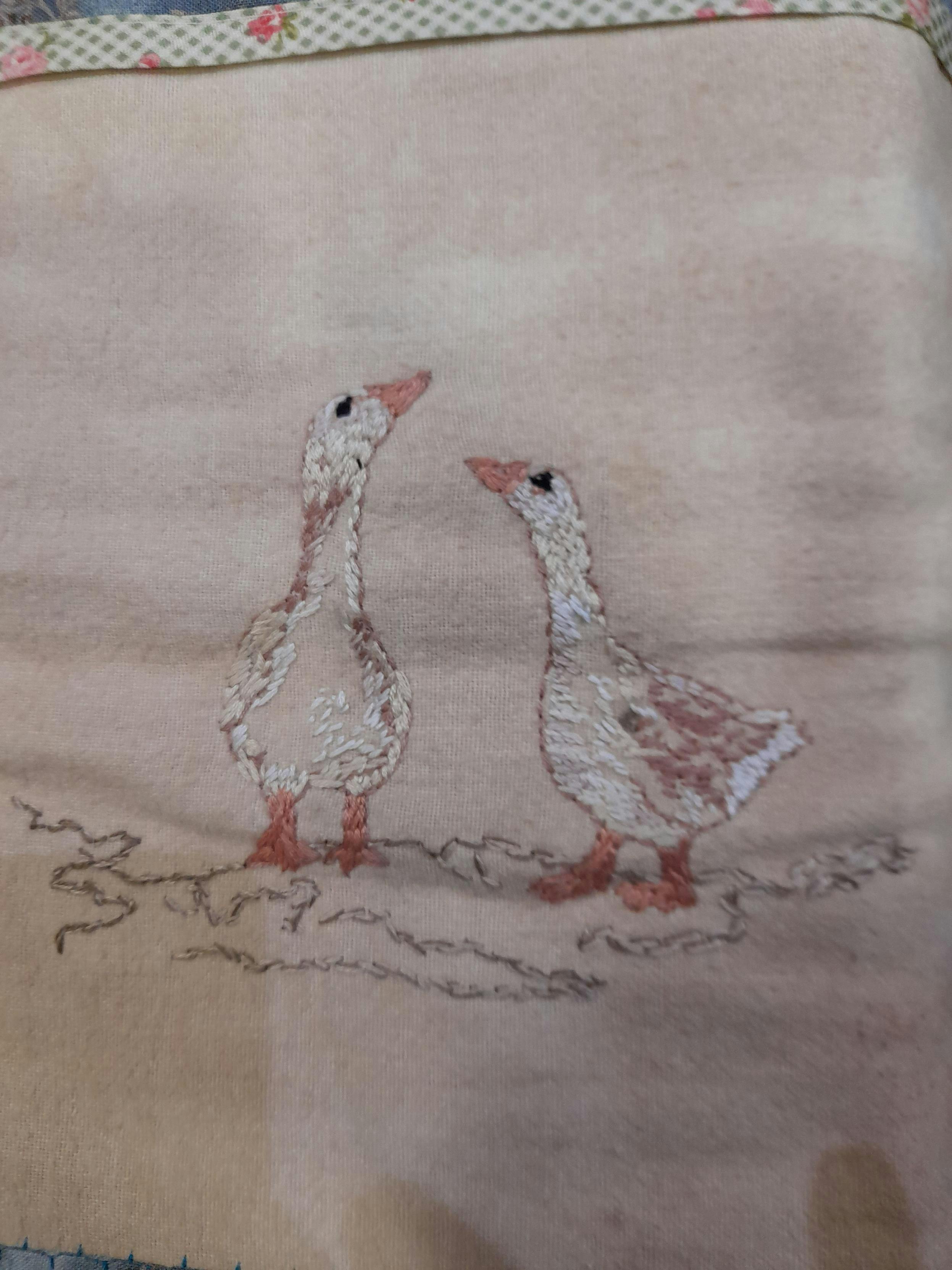 Two embroidered geese looking at one another, mostly creme white and some bright reddish brown. The fabric is dyed with black tea.