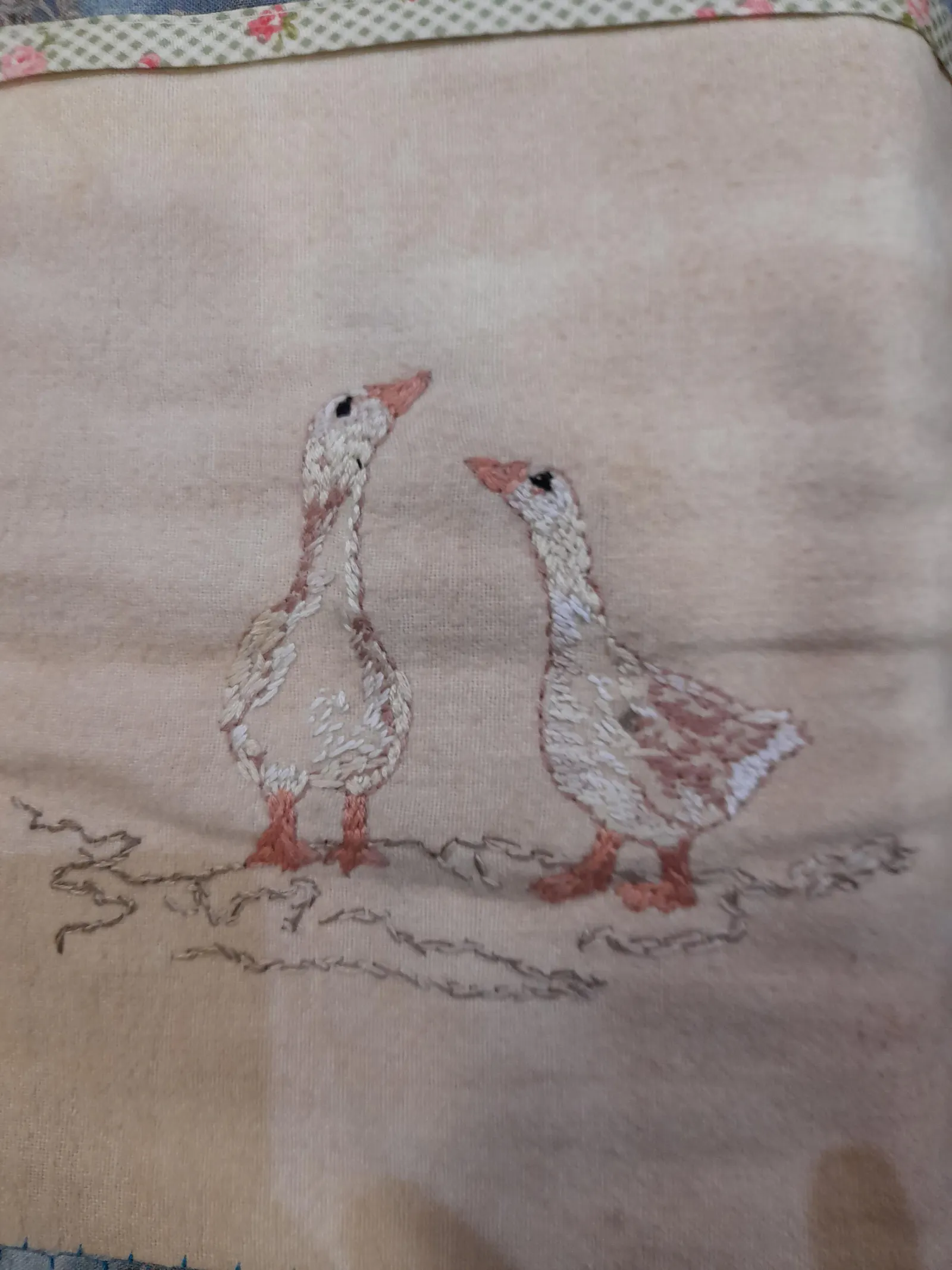 Two embroidered geese looking at one another, mostly creme white and some bright reddish brown. The fabric is dyed with black tea.