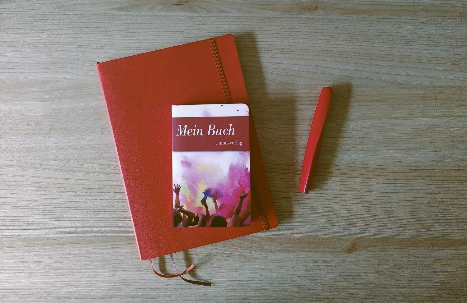 Ein rotes Notizbuch liegt auf dem Tisch, darauf ein kleineres Buch mit dem Titel "Mein Buch", daneben ein roter Füller.