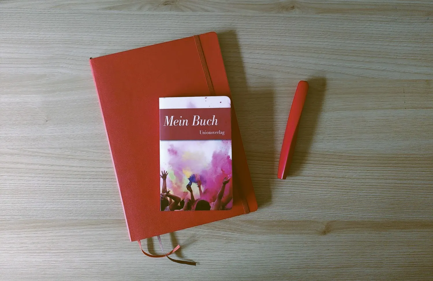 Ein rotes Notizbuch liegt auf dem Tisch, darauf ein kleineres Buch mit dem Titel "Mein Buch", daneben ein roter Füller.