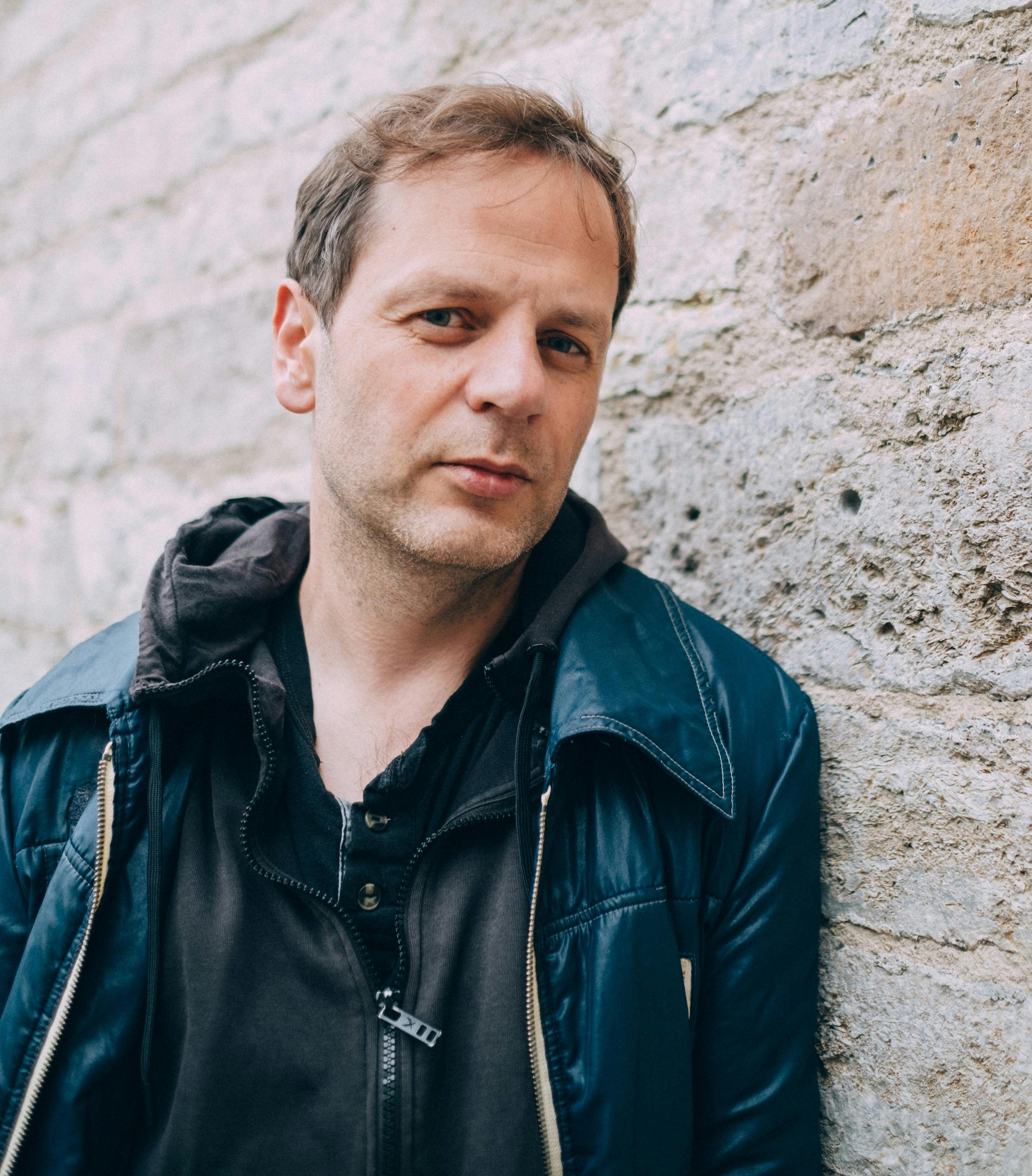 Ein mittelalter Mann mit braunen Haaren, schwarzem Pullover und blauer Lederjacke lehnt an einer Steinmauer.