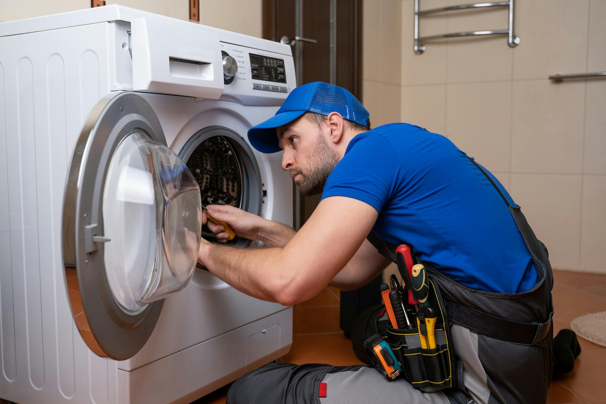 Dieses Foto zeigt einen Handwerker in blauer Arbeitskleidung, der vor einer weißen Waschmaschine kniet und sie repariert. Er hat eine Werkzeugtasche dabei.