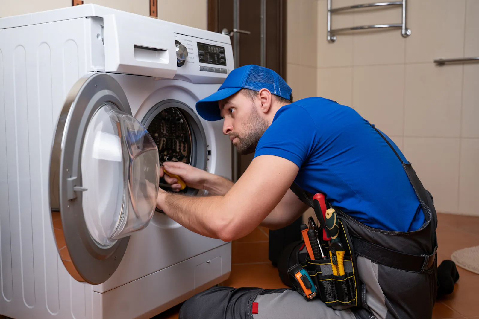 Dieses Foto zeigt einen Handwerker in blauer Arbeitskleidung, der vor einer weißen Waschmaschine kniet und sie repariert. Er hat eine Werkzeugtasche dabei.
