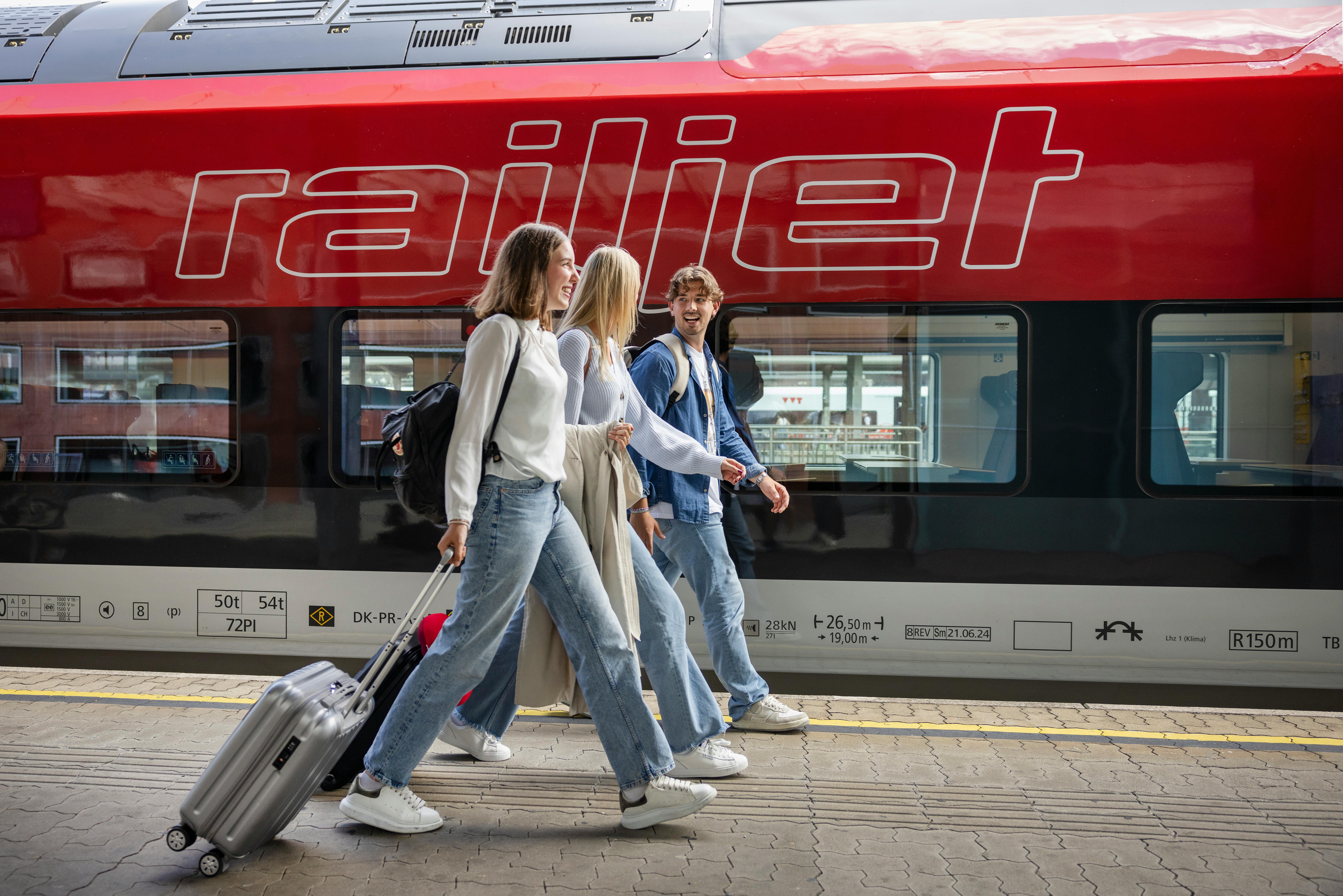 Dieses Foto zeigt drei junge Leute, die auf einem Bahnsteig vor einem roten Railjet der ÖBB entlanglaufen und einen Rollkoffer hinter sich herziehen.