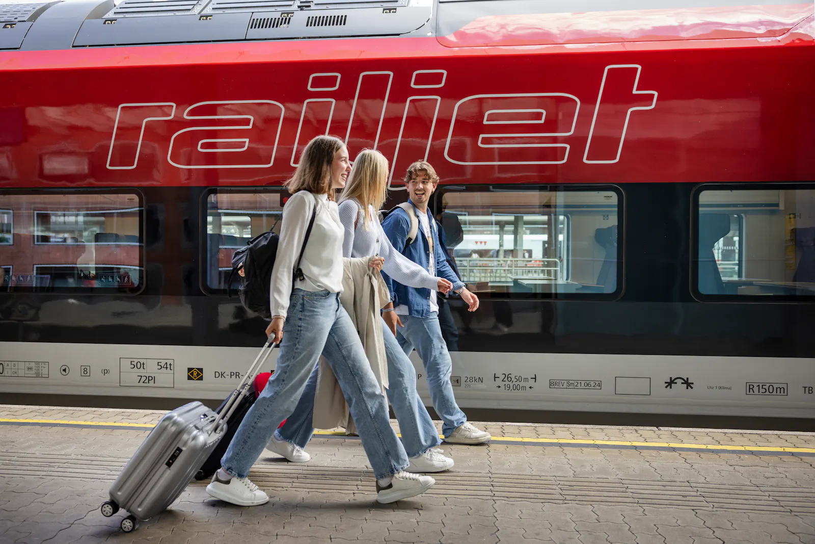 Dieses Foto zeigt drei junge Leute, die auf einem Bahnsteig vor einem roten Railjet der ÖBB entlanglaufen und einen Rollkoffer hinter sich herziehen.