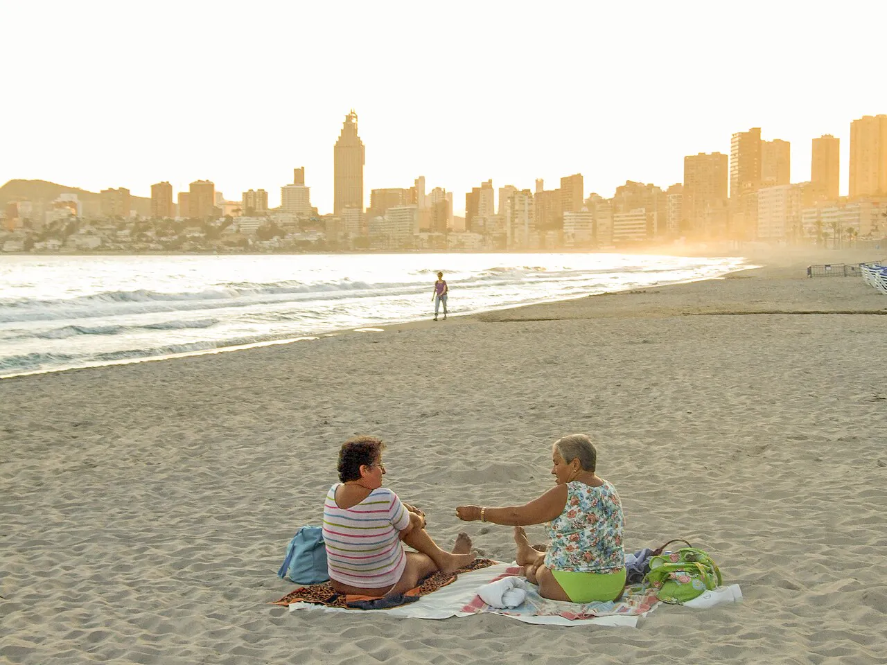 Zwei alte Frauen sitzen auf einem Handtuch am Strand von Benidorm und unterhalten sich. Im Hintergrund sieht man die Sykline des Ferienortes Benidorm
