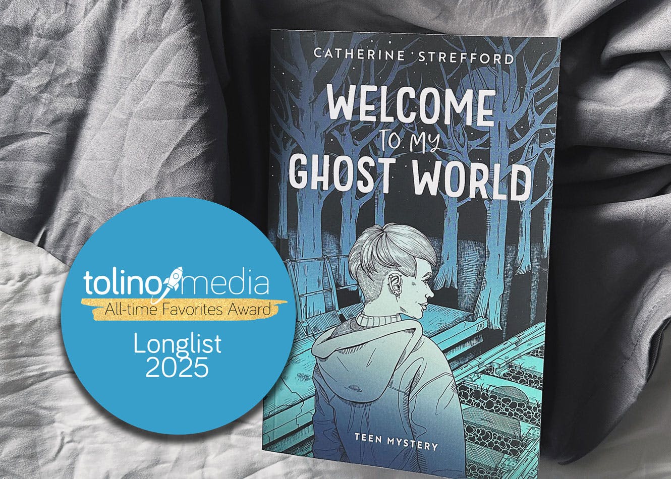 Das Taschenbuch von Welcome to my Ghost World auf einem ungemachten Bett. Auf dem Bild der Longlist-Badge vom tolino media All-Time Favorites Award.