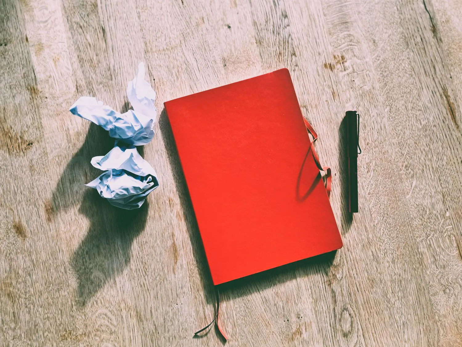 Ein rotes Notizbuch und ein FĂŒller liegen auf einer Tischplatte, daneben zwei zusammengeknĂŒllte Papierbögen.