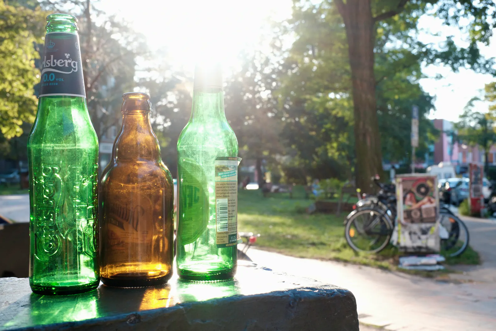 Drei leere Bierflaschen im Gegenlicht auf einer steinernen Erhöhung vor einem Park mit Fahrrädern.