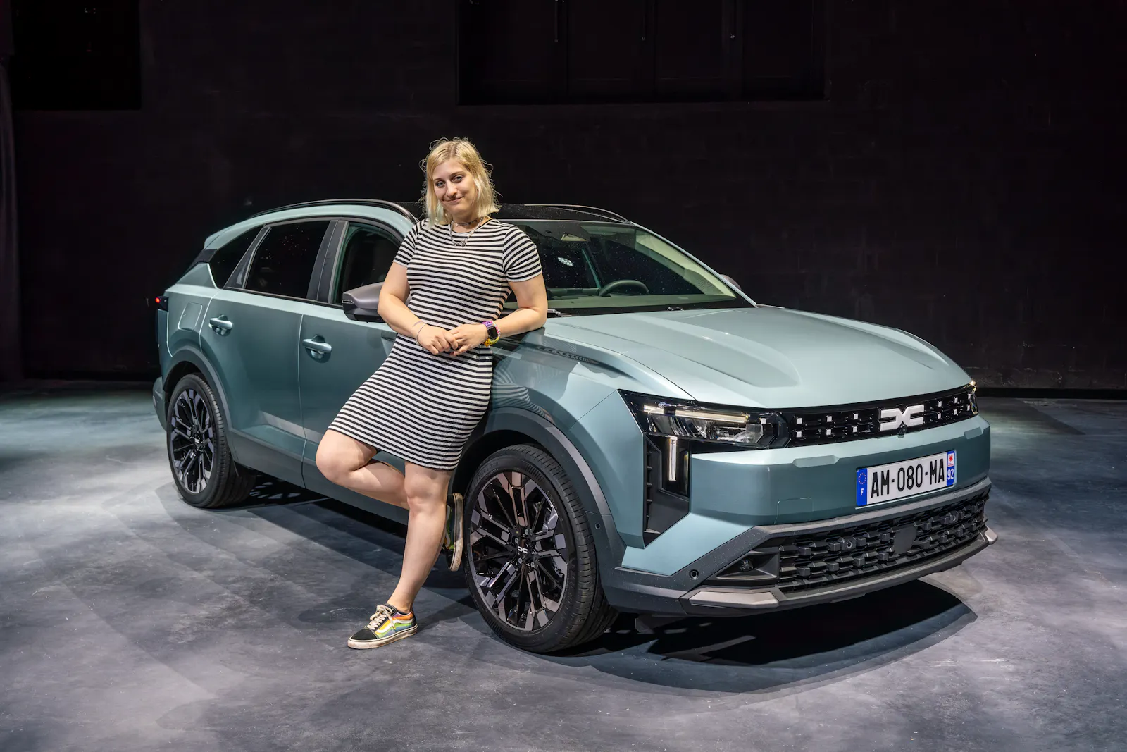 Carconny in einem gestreiften Kleid vor einem hellblauen Dacia Striker, einem Kombi Crossover. In einem Studio