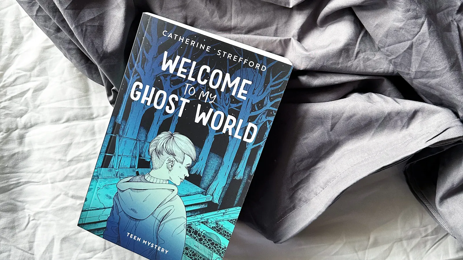 Das Buch "Welcome to my Ghost World", das auf zerknitterten weißen und grauen Laken liegt. Auf dem Cover ist eine Zeichnung der Protagonistin Lu zu sehen. Eine Rückenansicht, sie sieht in Richtung Wald, den sie von ihrer Position an einem Bahnsteig sehen kann. Sie hat kurze Haare und trägt eine Kaputzenjacke. Die Farben des Covers sind blau und türkis.