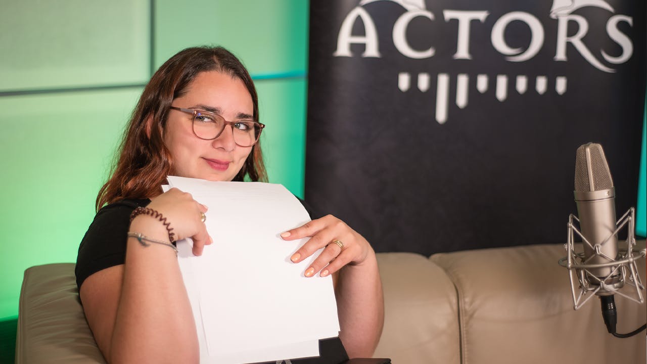 Aylin hält einige Zettel für die Planung des Drehwochenendes in der Hand. Sie sitzt auf einem Sofa, vor ihr steht ein Mikrofon und im Hintergrund sieht man ein Banner mit der Aufschrift „Dice Actors“ vor einem grünen Hintergrund.