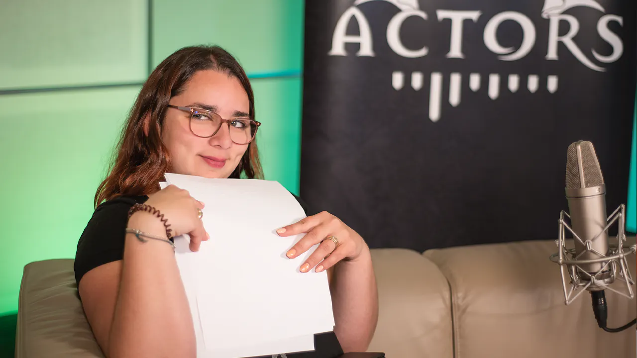 Aylin hält einige Zettel für die Planung des Drehwochenendes in der Hand. Sie sitzt auf einem Sofa, vor ihr steht ein Mikrofon und im Hintergrund sieht man ein Banner mit der Aufschrift „Dice Actors“ vor einem grünen Hintergrund.