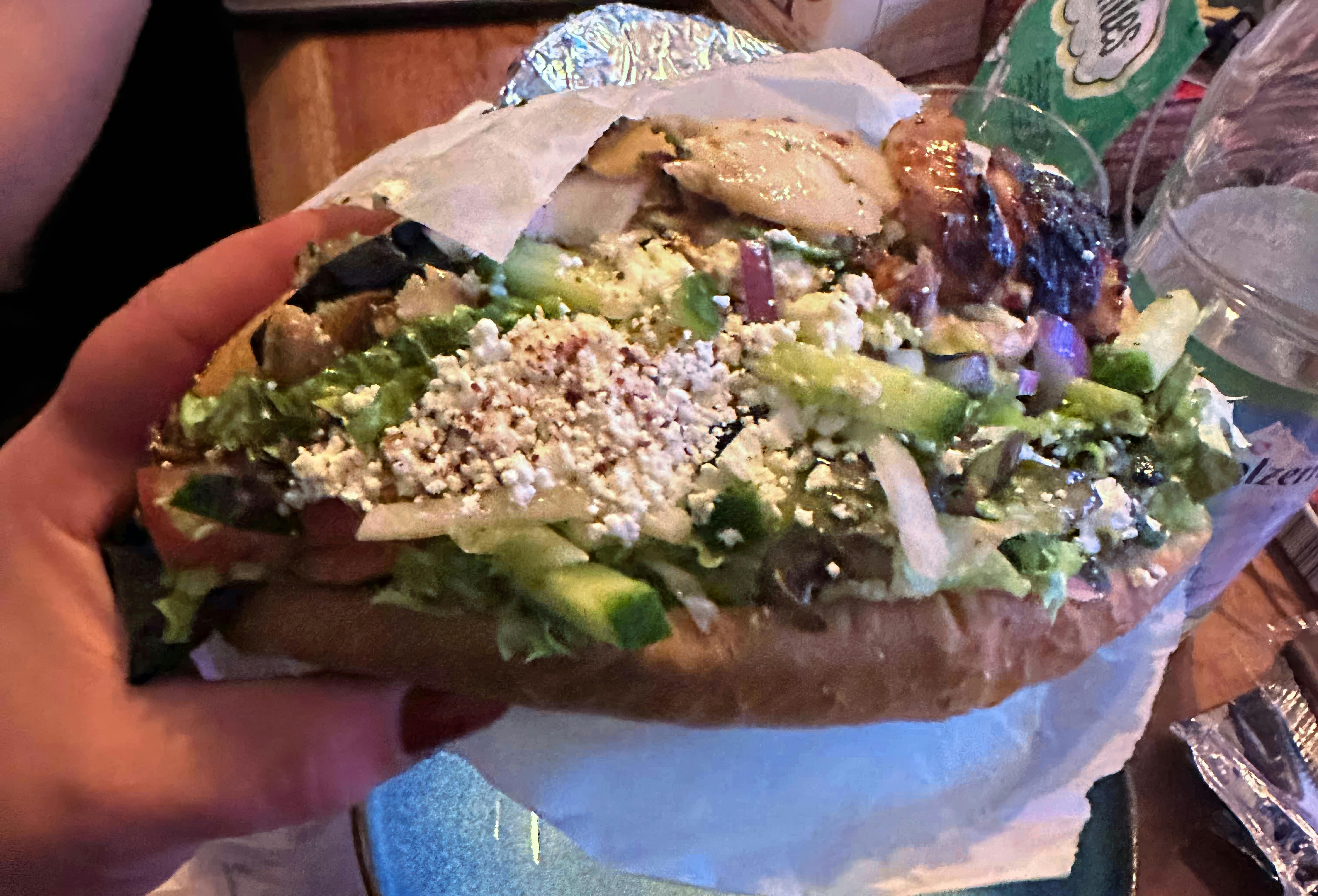 Aylin hält einen prall gefüllten Döner. Zu sehen sind viel frischer Salat, Gurke, Tomate, rote Zwiebeln, Kräuter und eine große Portion zerbröselter Käse. Oben ragt gegrilltes Fleisch heraus. Der Döner liegt in einem aufgeklappten Fladenbrot und ist in Papier gewickelt.
