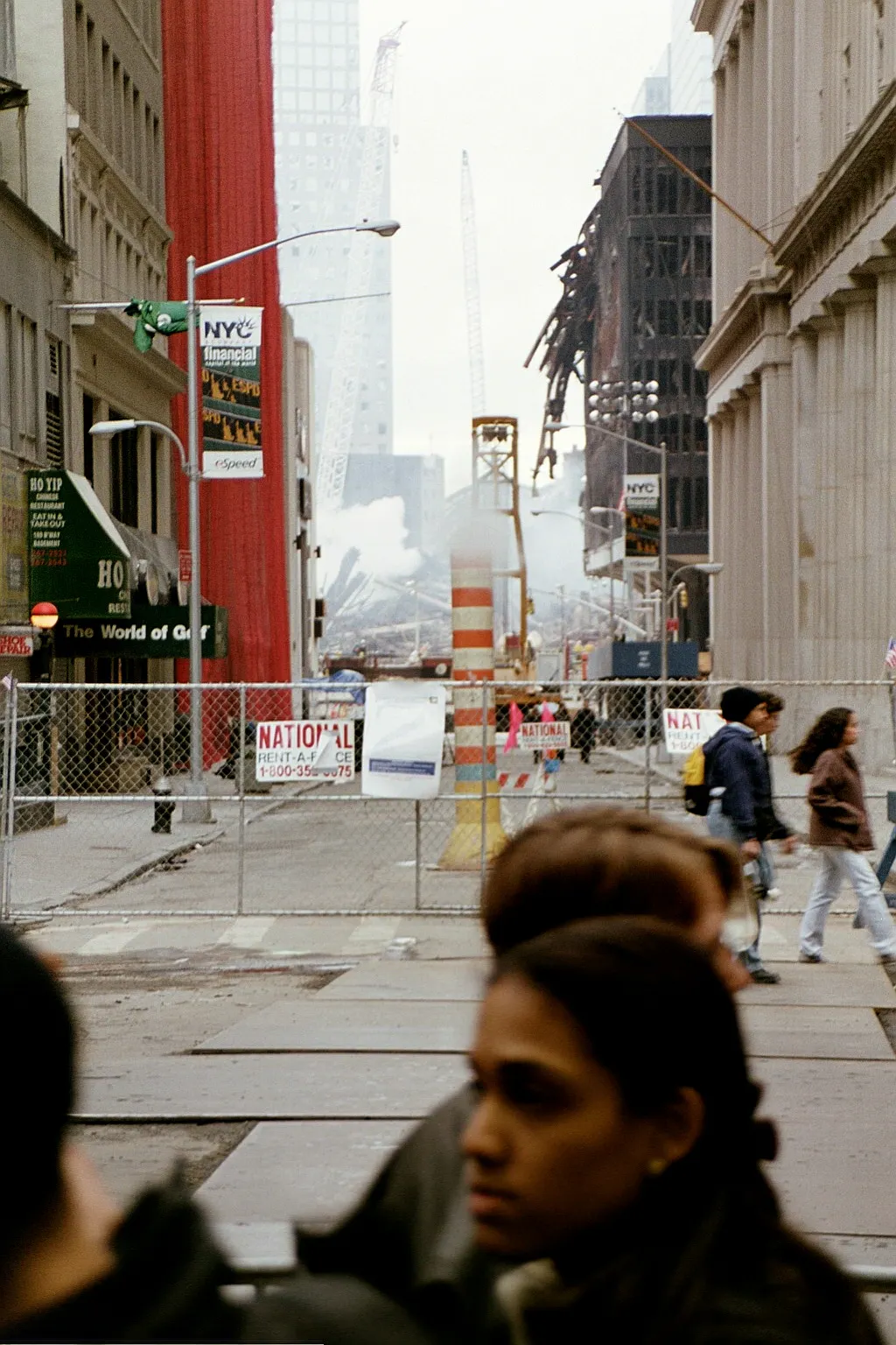 Lower Manhattan, Passant*innen vor einer Absperrung, am Ende einer Straße sieht man Trümmer des World Trade Centers, Rauch hängt in der Luft.
