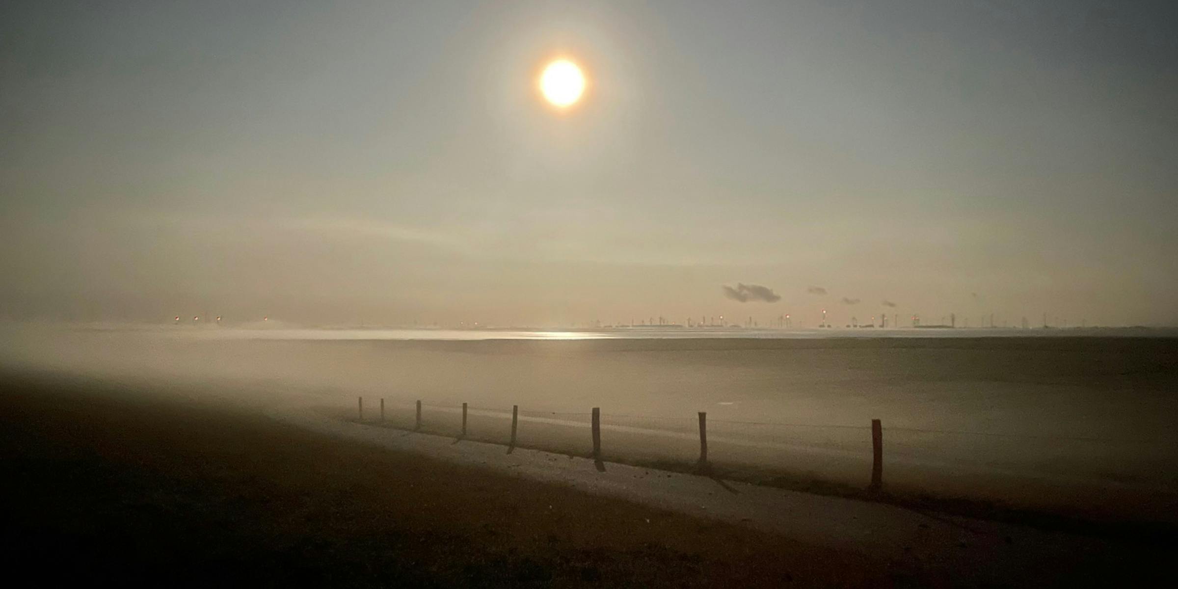 Ein heller Mond über einer Küstenlandschaft, im Vordergrund ein Zaun, links zieht Bodennebel heran.