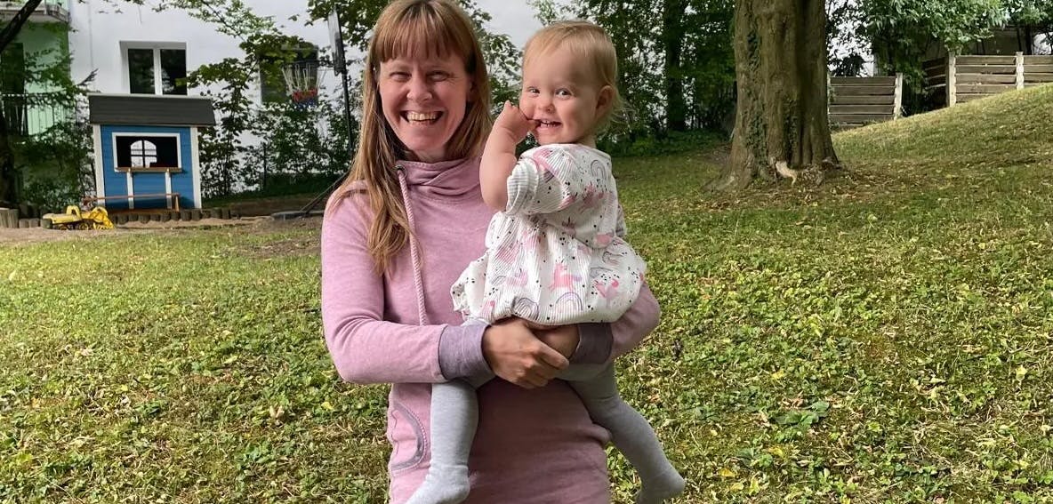 Das Portrait von Sandra Hauser, einer jungen lächelden Theologin mit Kind auf dem Arm vor ihrem Haus im Grünen.