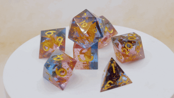 Sieben Würfel aus durchsichtigem Kunstharz in einem warmen Farbverlauf aus Rosa, Blau und Bernsteingelb mit goldenen Zahlen, dekorativ auf einem weißen Drehteller angeordnet.