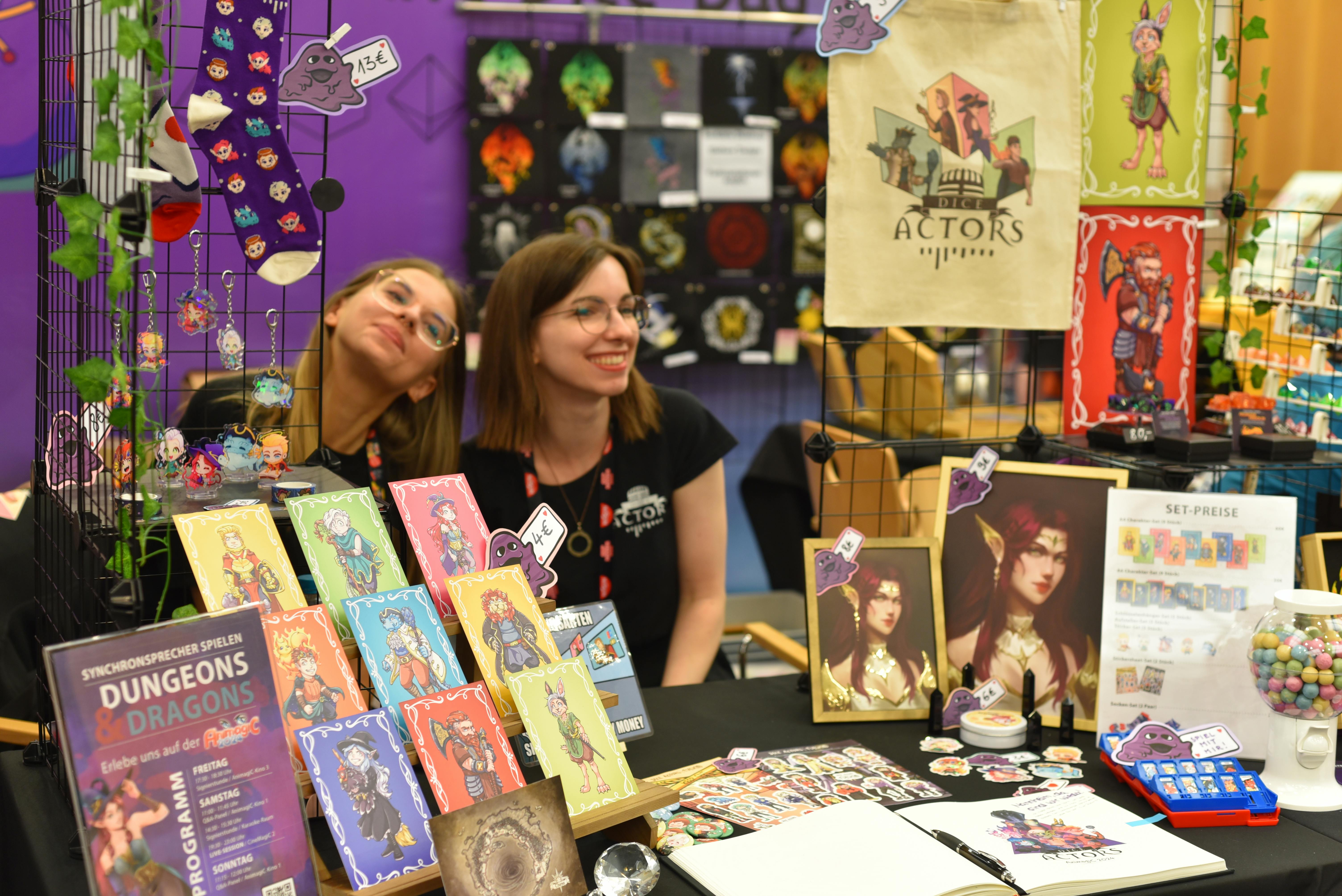 Susa und Franzi sitzen lächelnd hinter einem bunten Merchandise-Stand der „Dice Actors" auf der AnimagiC 2024. Auf dem Tisch liegen Prints, Sticker, Buttons und weitere schöne Artikel.