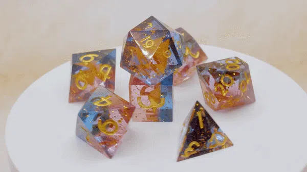 Sieben Würfel aus durchsichtigem Kunstharz in einem warmen Farbverlauf aus Rosa, Blau und Bernsteingelb mit goldenen Zahlen, dekorativ auf einem weißen Drehteller angeordnet.