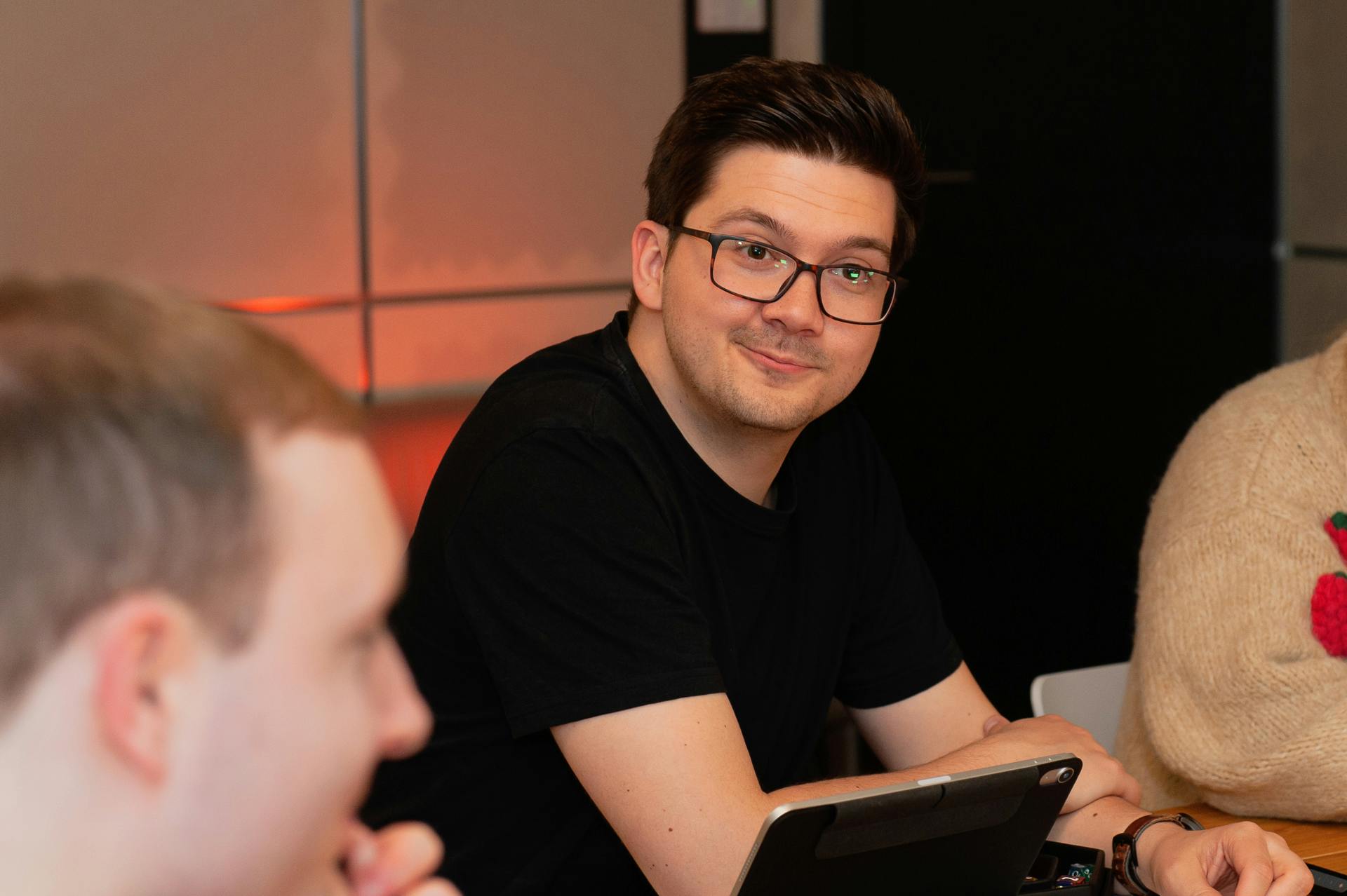 Patze sitzt am Spieltisch mit einem Tablet vor sich und lächelt schief. Im Vordergrund ist Kelly unscharf zu sehen, im Hintergrund warmes orangefarbenes Licht.
