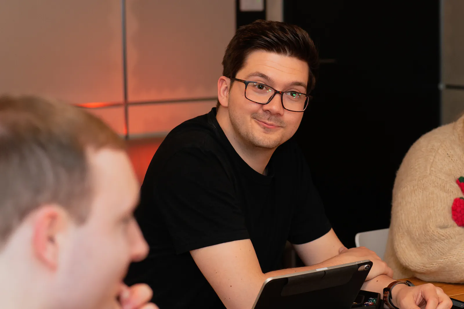 Patze sitzt am Spieltisch mit einem Tablet vor sich und lächelt schief. Im Vordergrund ist Kelly unscharf zu sehen, im Hintergrund warmes orangefarbenes Licht.