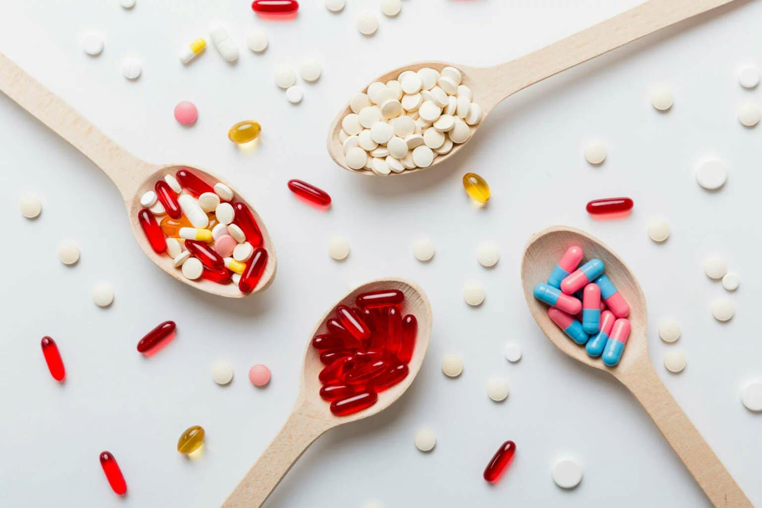 Mehrere Löffel mit unterschiedlich gefärbten Tabletten und Kapseln auf hellem Hintergrund.
